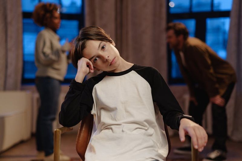 A young boy in a black and white shirt looks sad as his parents argue in the background.