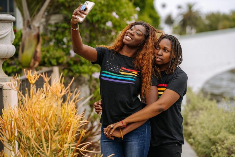 Ein verliebtes Paar macht ein Selfie und genießt den Moment in LGBT-Pride-Shirts vor einer malerischen Kulisse.