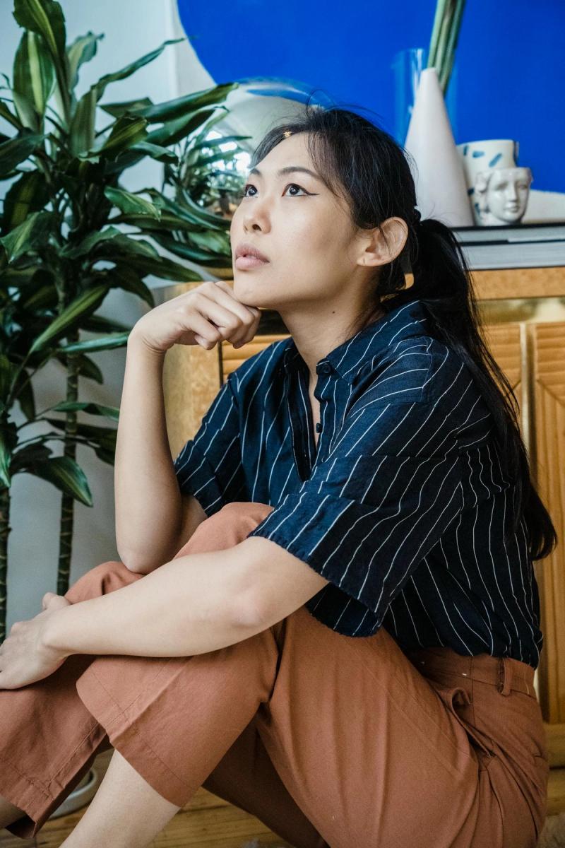 Asian woman sitting thoughtfully indoors beside indoor plants.