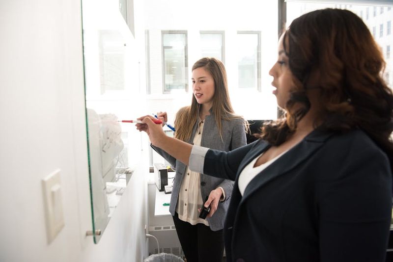 Two women engaging in teamwork by writing on a whiteboard in a bright office setting.