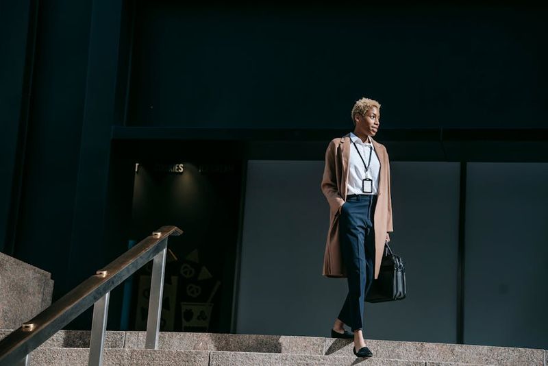 From below full body positive young female entrepreneur in stylish brown coat with hand in pocket and bag walking down stairs near office building