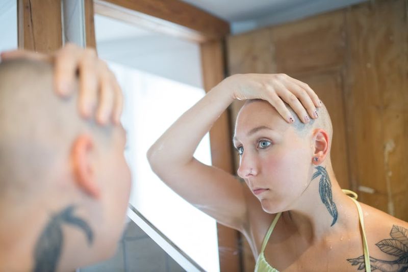Bald woman examines her reflection in an indoor setting, highlighting tattoos and introspection.