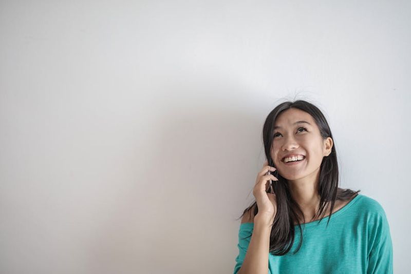 Smiling Asian woman talking on a smartphone indoors, looking upwards with a joyful expression.