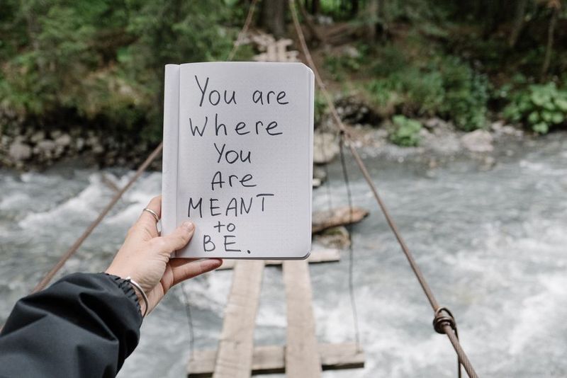 Nahaufnahme eines Notizbuchs mit einer inspirierenden Botschaft, gehalten über einem Fluss mit einer Holzbrücke.
