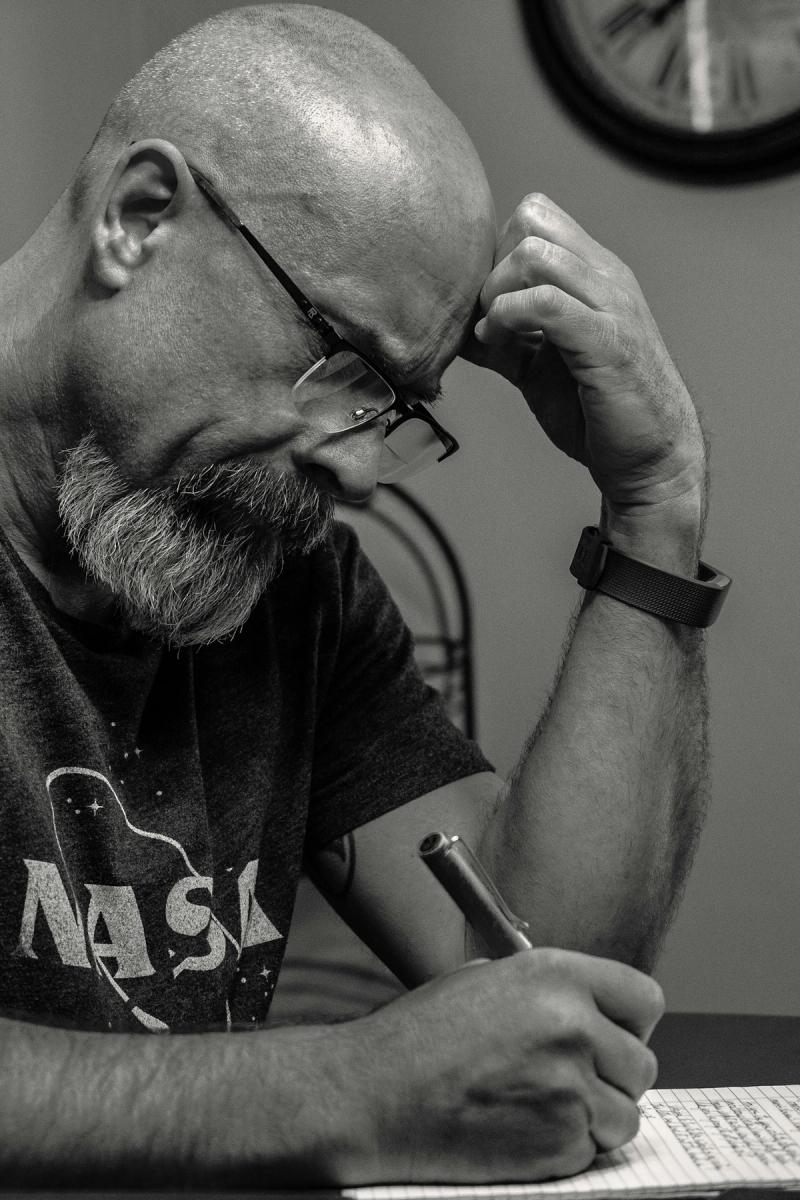 Black and white image of a thoughtful senior man writing indoors, wearing glasses and a NASA shirt.