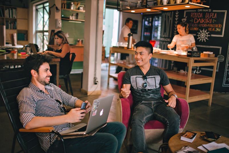 Dois homens desfrutando de uma conversa descontraída com laptops em um aconchegante café brasileiro.