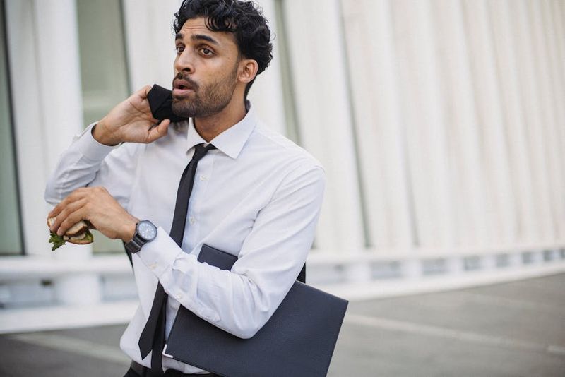 A businessman in corporate attire multitasking with a sandwich and phone outdoors.