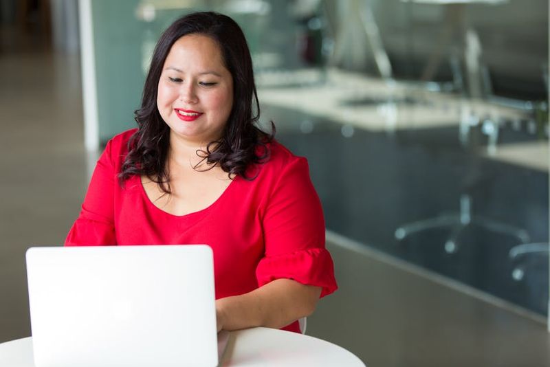 Asian woman smiling while working on a laptop in a modern office environment.