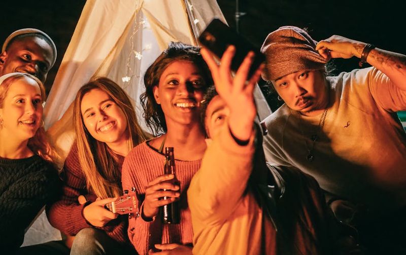 A joyful group of friends takes a selfie around a tent, sharing laughter and warmth at a nighttime gathering.