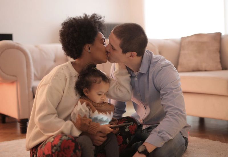 A loving couple kisses while sitting with their child in a warm living room setting.