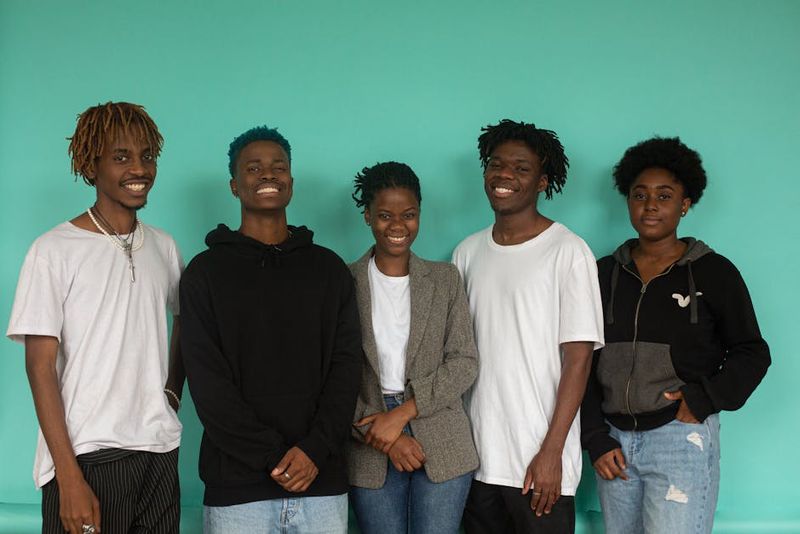 A diverse group of five young adults smiling against a teal background in a studio setting.