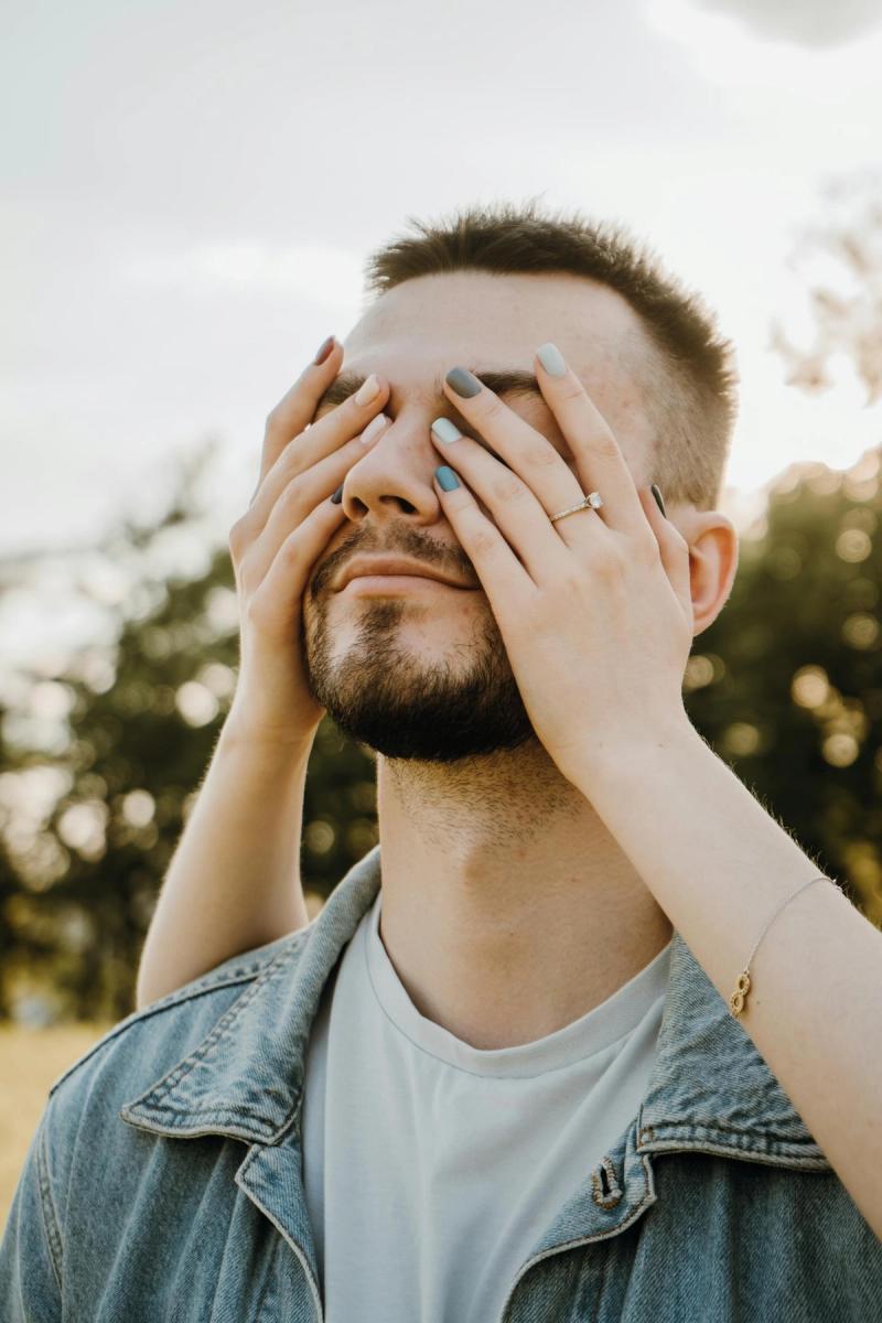 Nahaufnahme eines lächelnden Mannes im Freien, der sich spielerisch die Augen zuhält. Warmherziger und ungezwungener Moment.