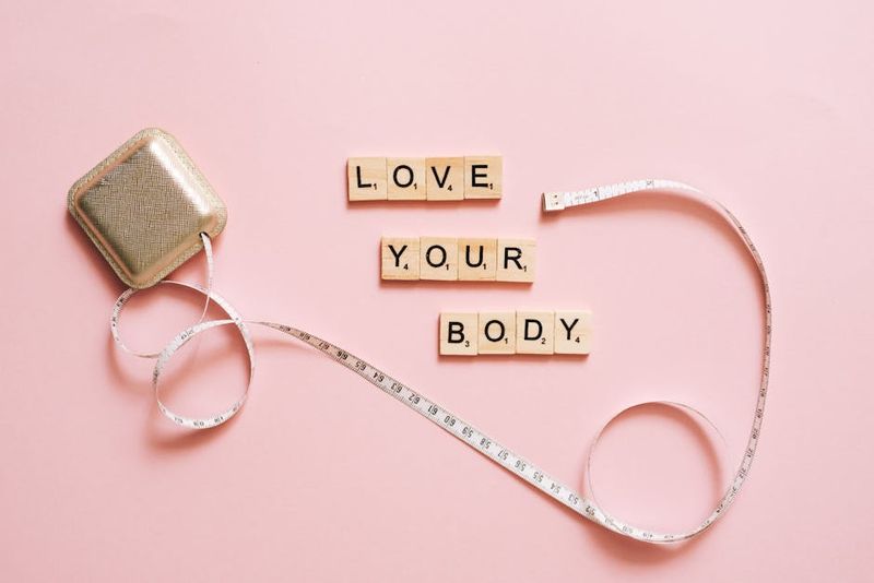 Scrabble letters spelling 'Love Your Body' with a tape measure on a pink backdrop.