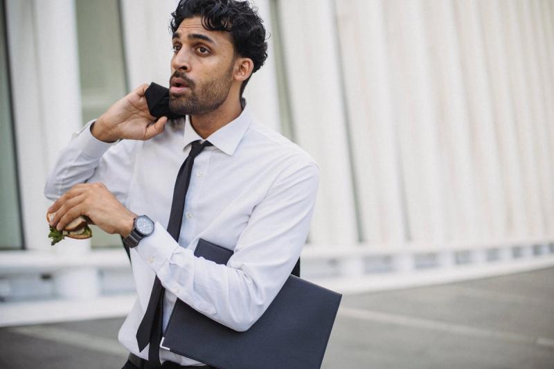 Un uomo d'affari in abbigliamento formale che fa multitasking con un panino e un telefono all'aperto.