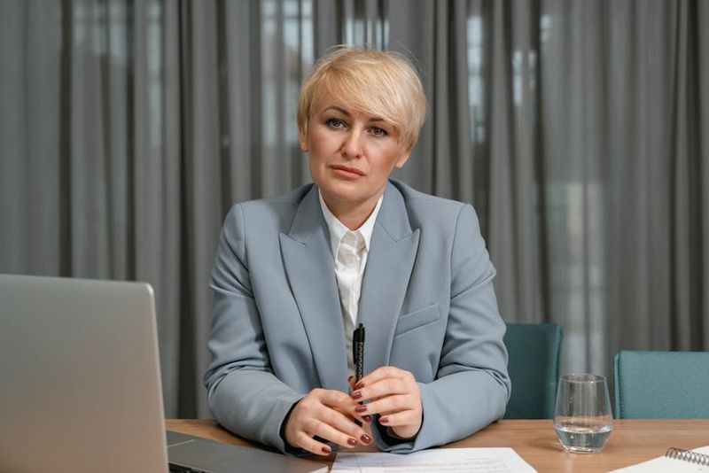 Una donna d'affari sicura di sé in abito blu è seduta alla scrivania con un laptop, una penna e dei documenti in un ambiente da ufficio.