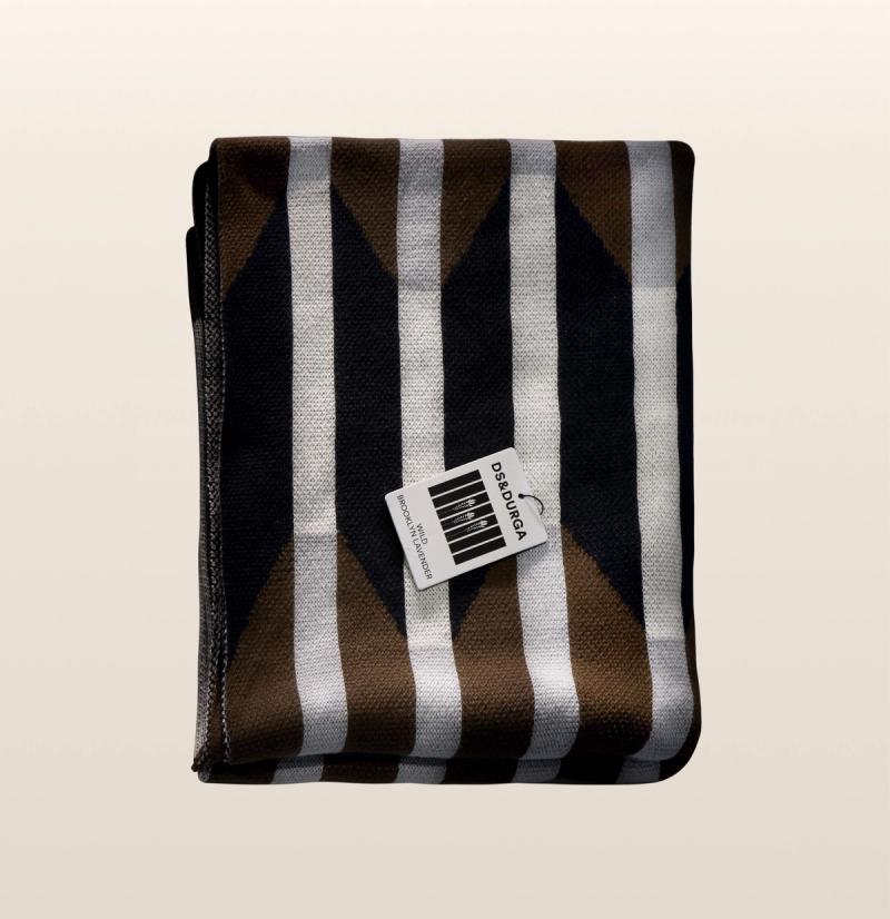 A brown, black, white, and gray woven cotton blanket with a geometric pattern, folded into a rectangle, with a DS & Durga Wild Brooklyn Lavender auto fragrance resting on top of it.