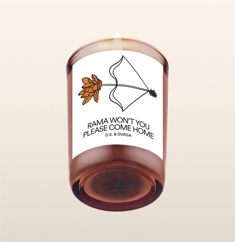 A lit scented candle featuring an illustration of a flower and a kite, and the text "rama, won't you please come home" by d.s. & durga.
