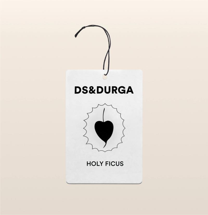 A hanging auto fragrance with the inscription "ds&durga" at the top and the name "holy ficus" below an emblem featuring a leaf silhouette inside a jagged circle. 