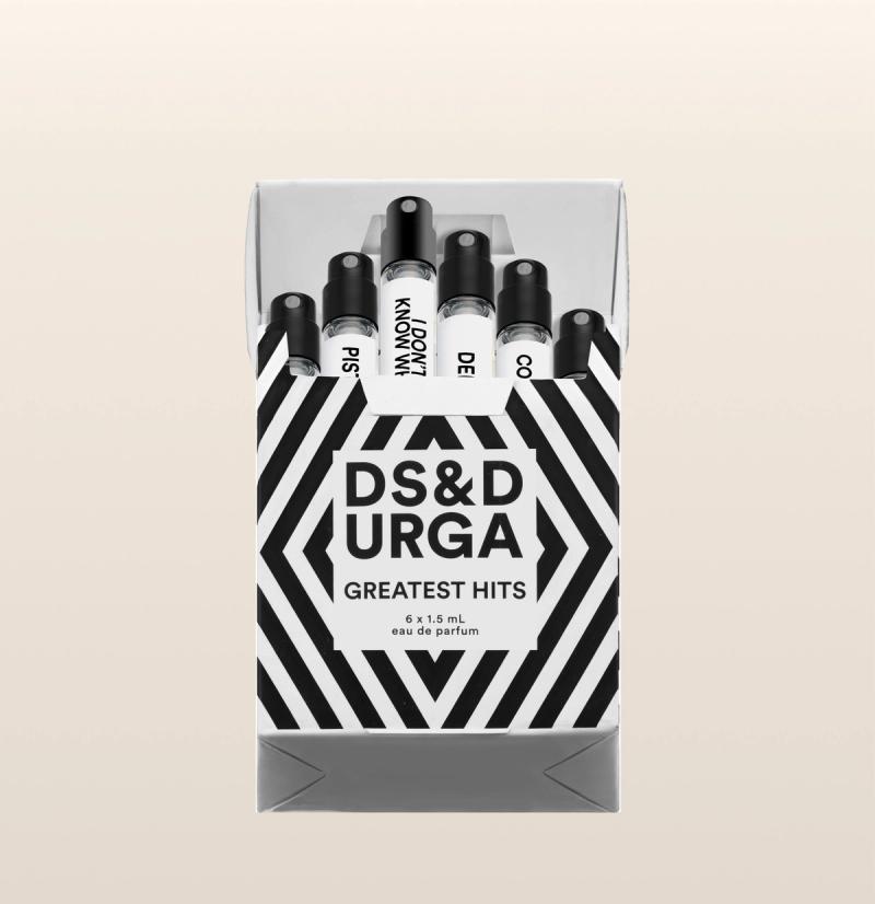 A black and white striped box labeled "DS & DURGA GREATEST HITS" holds six small perfume vials, with their labels and black caps visible above the open box top. The background is light beige.