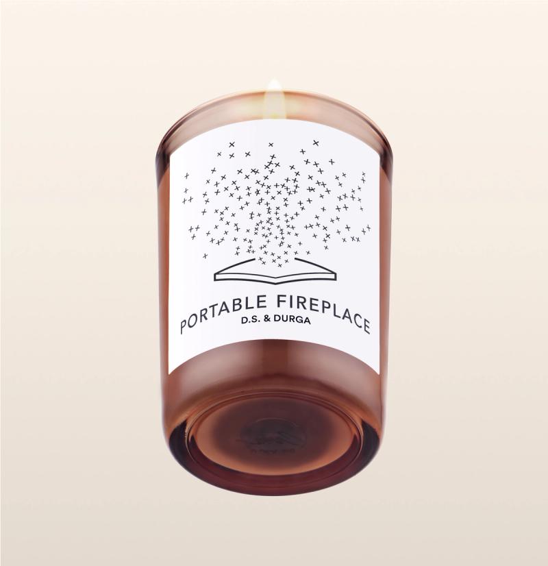 A lit illuminated scented candle with a whimsical "portable fireplace" label by d.s. &amp; durga, casting a warm glow through amber-colored glass.
