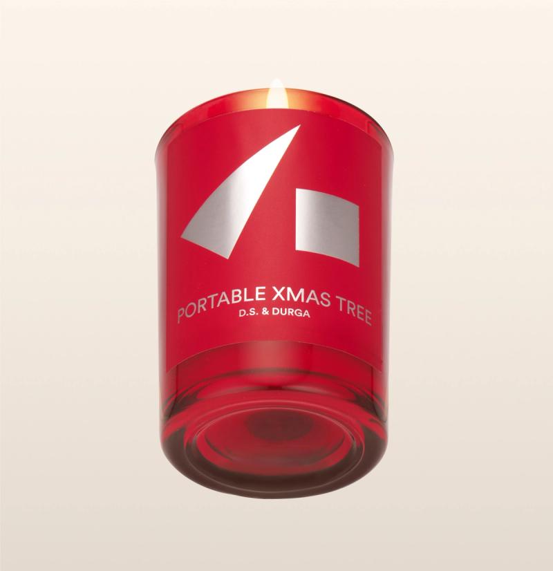 A red candle in a glass holder labeled "PORTABLE XMAS TREE D.S. &amp; DURGA," featuring minimalist silver geometric shapes on the front. The candle is lit against a plain, light background.