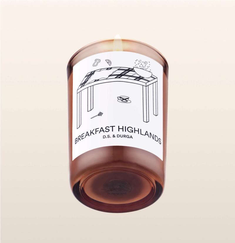A lit scented candle with a label that reads "breakfast highlands" by d.s. &amp; durga, featuring a simple illustration of a table with a coffee cup and a newspaper.