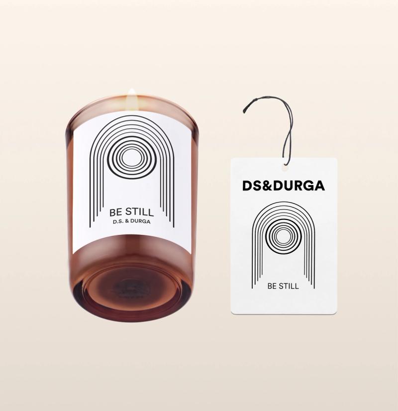 A brown candle in a glass holder with a minimalist black and white pattern and "Be Still D.S. & Durga" text. Beside it, a matching auto fragrance with similar design and text "DS & DURGA Be Still", attached by a black string.