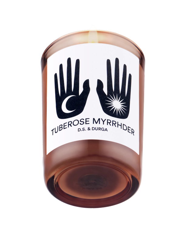 A lit scented candle with the label "tuberose myrrhdr" by d.s. & durga, featuring graphic black-and-white hand symbols on the label.