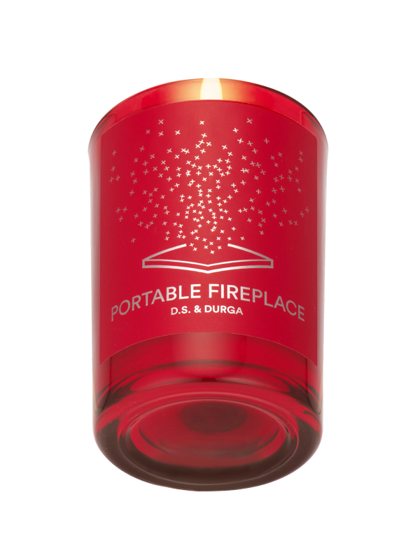 A red glass candle labeled "PORTABLE FIREPLACE D.S. &amp; DURGA," with a flame burning and a pattern of small stars above an open book design on the front.