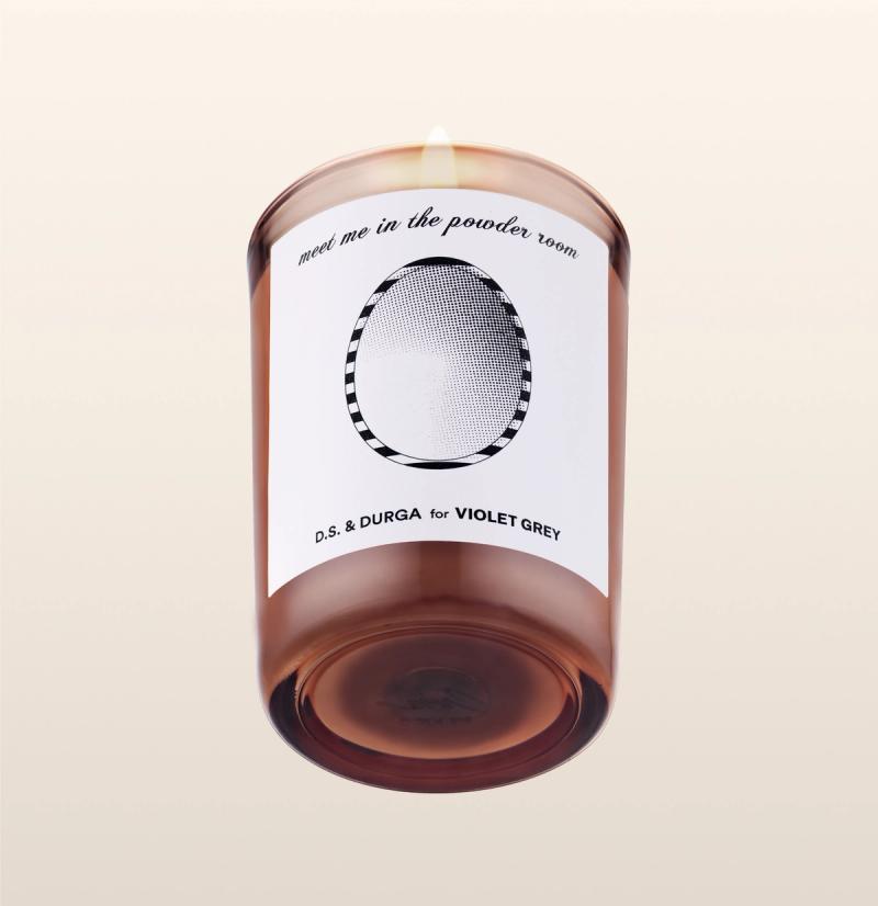 A brown glass candle with a lit flame, labeled "meet me in the powder room" above a black-and-white oval design. The label also reads "D.S. & DURGA for VIOLET GREY." The background is plain and light-colored.