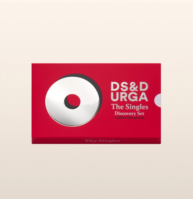 Rectangular red box with a white circular design on the left and text on the right that reads “DS&amp;D URGA The Singles Discovery Set Coffret Découverte.” The background is a light beige gradient.