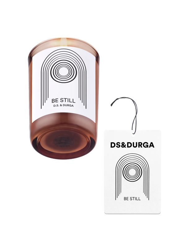 A brown candle in a glass holder with a minimalist black and white pattern and "Be Still D.S. & Durga" text. Beside it, a matching auto fragrance with similar design and text "DS & DURGA Be Still", attached by a black string.