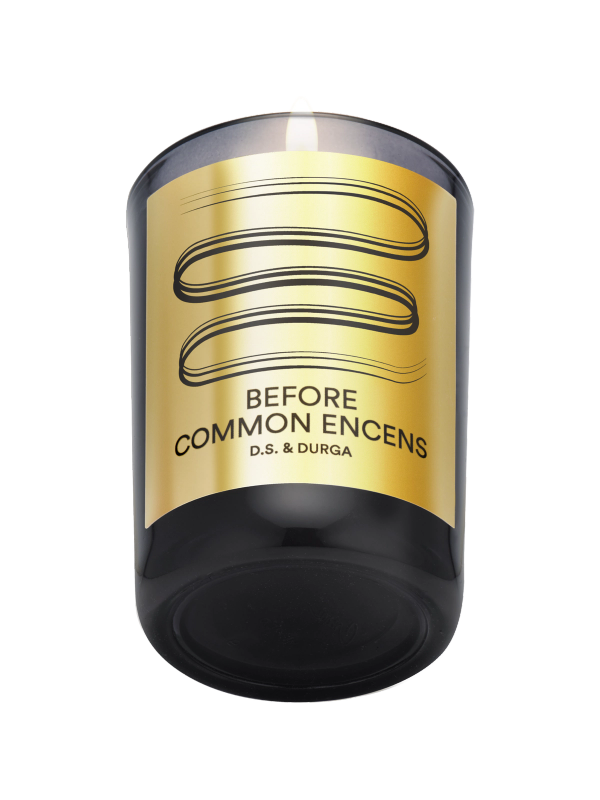 A black candle with a gold label reading "Before Common Encens" and "D.S. & Durga." The label features a simple abstract line design above the text.