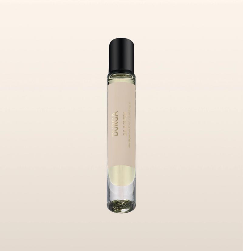 A clear glass perfume roller bottle with a black cap, containing pale yellow liquid. The word "DURGA" and additional text appear in gold on the bottle. The background is a light beige gradient.