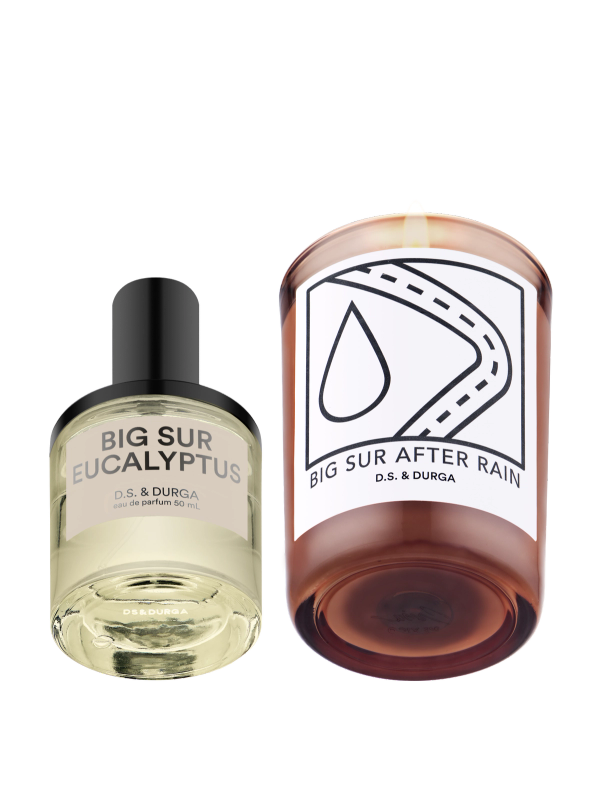 A clear bottle of D.S. & Durga Big Sur Eucalyptus eau de parfum stands next to a brown glass candle labeled "Big Sur After Rain" with a graphic of a water droplet and winding road on the candle.