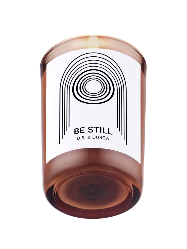 A lit scented candle with label reading "be still"by d.s. &amp; durga, showcasing minimalist design on a transparent background.