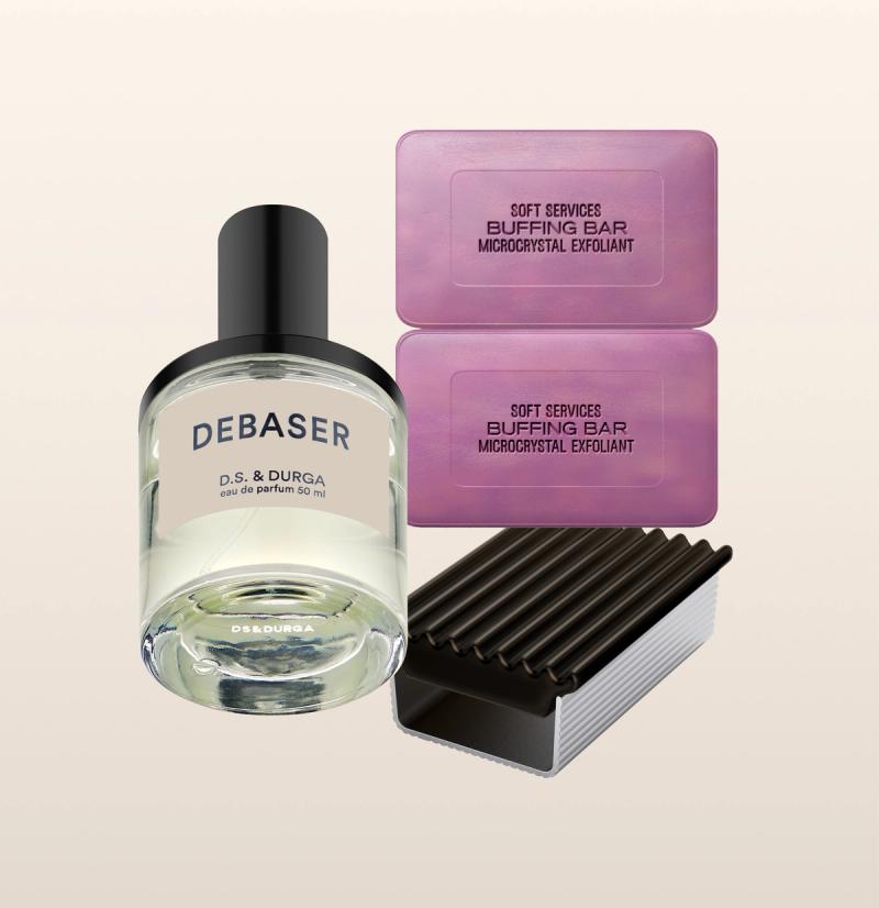 An arrangement of D.S. & Durga Debaser eau de parfum bottle and two Soft Services buffing bar microcrystal exfoliants. The perfume bottle is in the foreground, while the two purple buffing bars and their dark purple ridged holder are in the background.