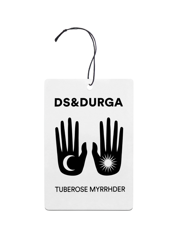 A hanging auto fragrance bold typography showcasing the brand "ds & durga" and the product name "tuberose myrrhder". the label features two stylized, symmetrical icons of an open hand, one with an eye in the palm and the other with a radiating star or burst.