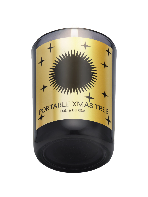 A lit candle in a dark glass holder with a gold label. The label features a black sunburst design and small stars, with the text "Portable Xmas Tree" and "D.S. & Durga."