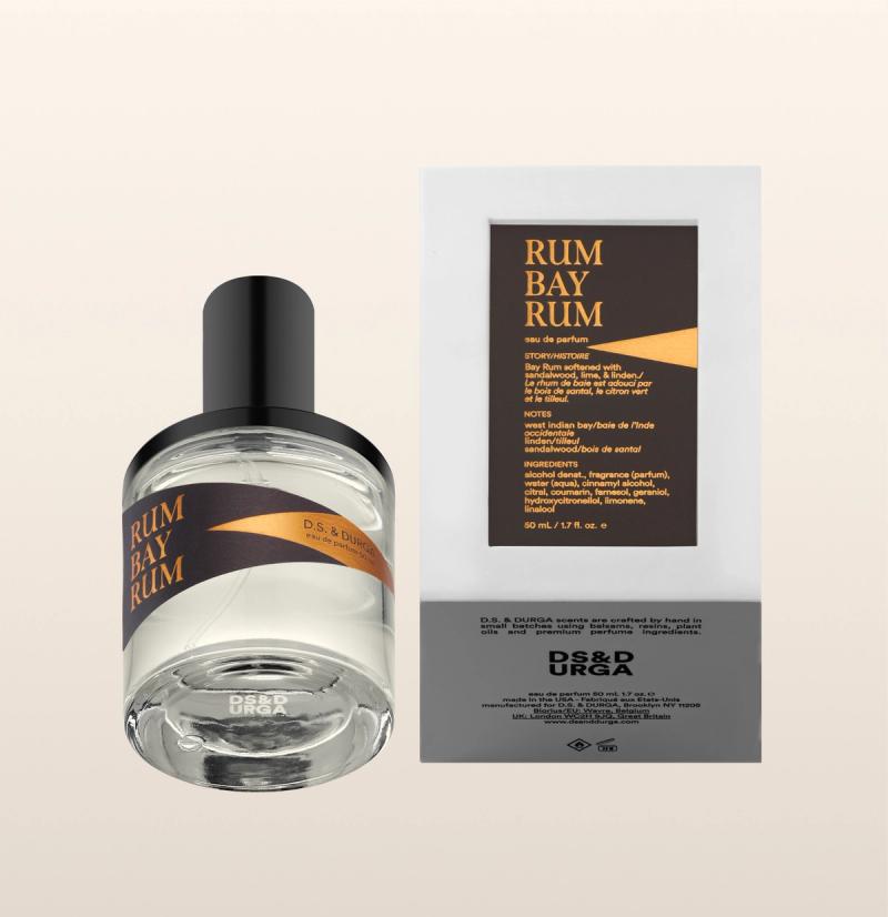 A clear perfume bottle labeled "RUM BAY RUM" lies next to its white rectangular box, which has a matching label with yellow text and a black and brown design. The brand "DS & DURGA" is visible on both.
