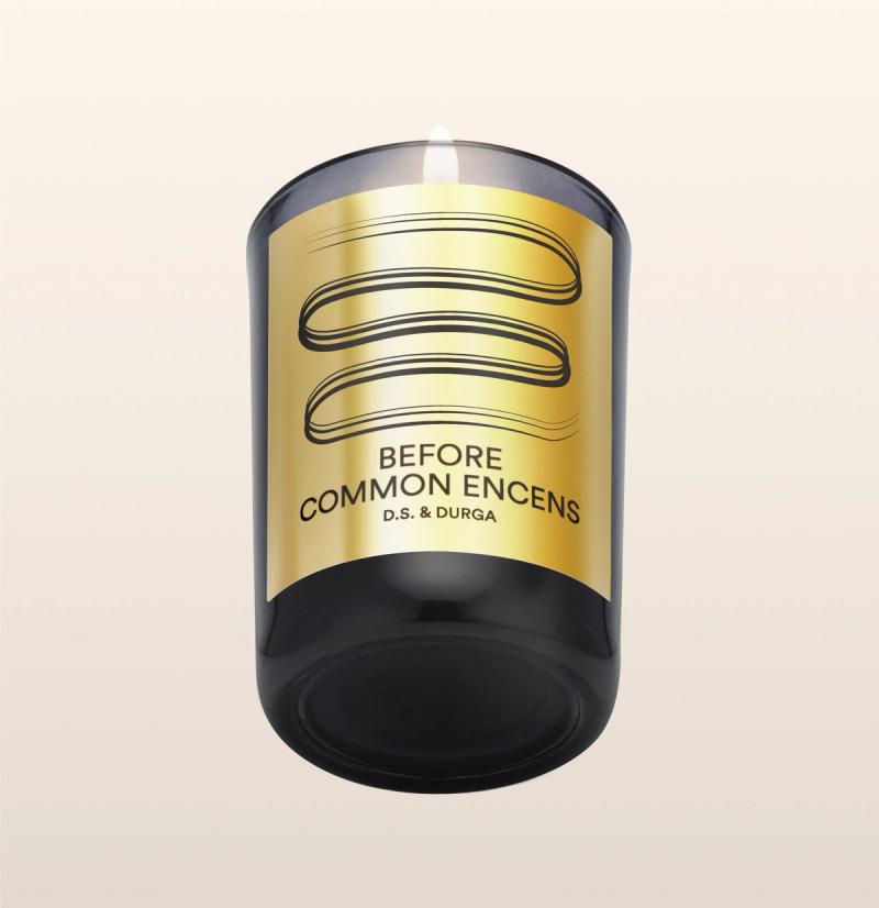 A lit candle in a black glass holder with a metallic gold label. The label features three wavy lines above the text "BEFORE COMMON ENCENS D.S. & DURGA." The background is a soft gradient from cream to light beige.