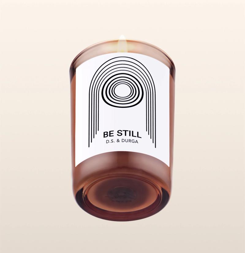 A lit scented candle with label reading "be still" by d.s. &amp; durga, showcasing minimalist design on a transparent background.