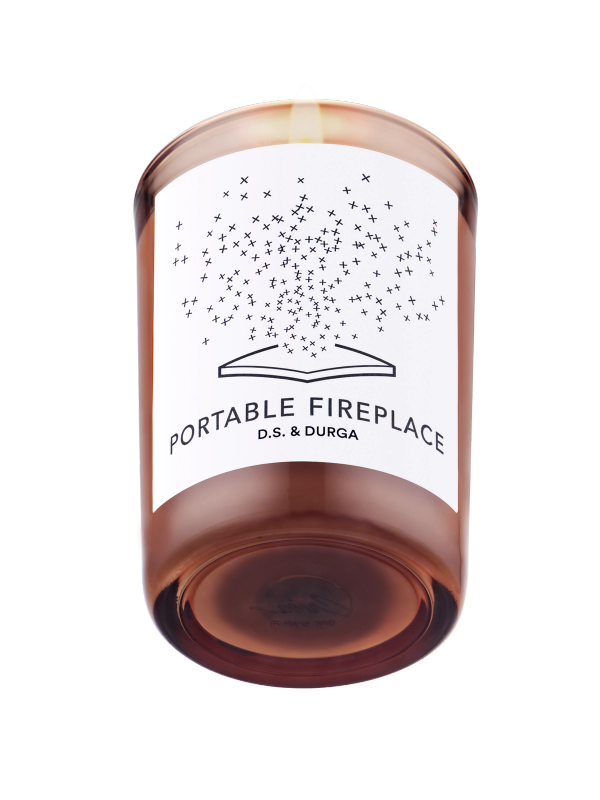 A lit illuminated scented candle with a whimsical "portable fireplace" label by d.s. &amp; durga, casting a warm glow through amber-colored glass.