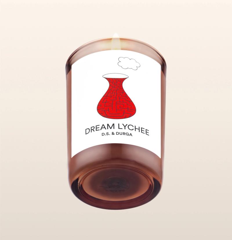 A lit scented candle with a warm, amber hue labeled "dream lychee" by d.s. &amp; durga, featuring an artistic red sketch of a traditional vase on its label.