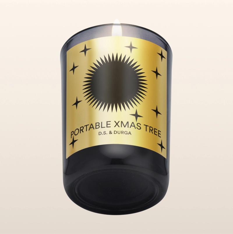 A lit candle in a dark glass holder with a gold label. The label features a black sunburst design and small stars, with the text "Portable Xmas Tree" and "D.S. & Durga" on a beige gradient background.