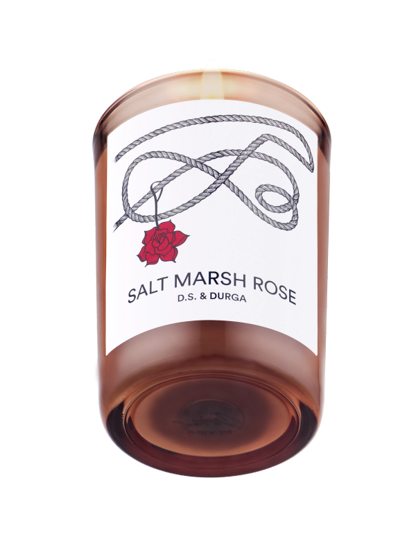 A lit scented candle with the label "salt marsh rose" by d.s. &amp; durga, featuring a simplistic design with a rose illustration against a clear background, casting a warm glow through its amber-colored wax.