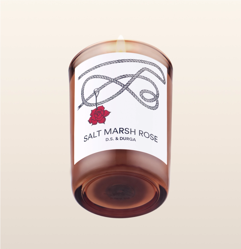 A lit scented candle with the label "salt marsh rose" by d.s. &amp; durga, featuring a simplistic design with a rose illustration against a clear background, casting a warm glow through its amber-colored wax.
