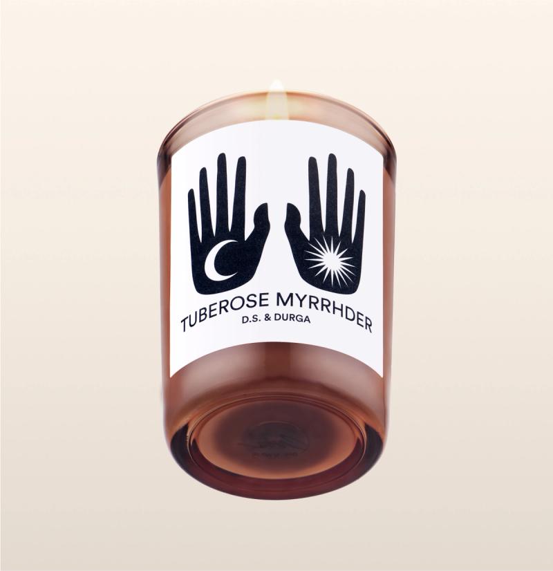 A lit scented candle with the label "tuberose myrrhdr" by d.s. & durga, featuring graphic black-and-white hand symbols on the label.