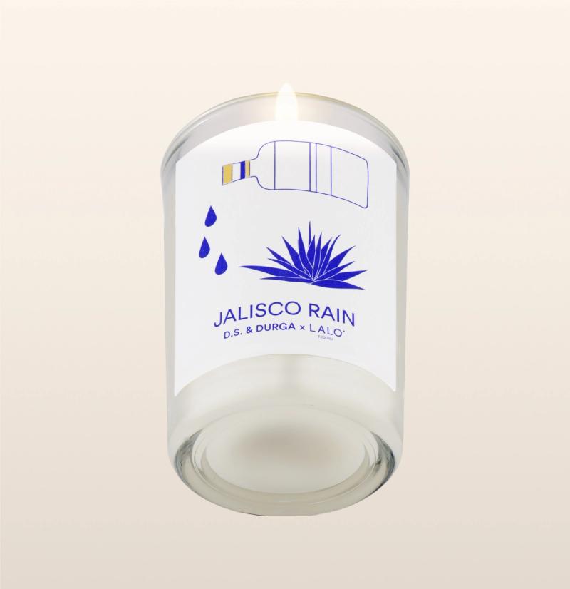 A lit candle in a clear glass holder is shown against a light background. The label reads "JALISCO RAIN D.S. & DURGA x LALO" in blue text, with illustrations of a bottle pouring liquid, water drops, and an agave plant.