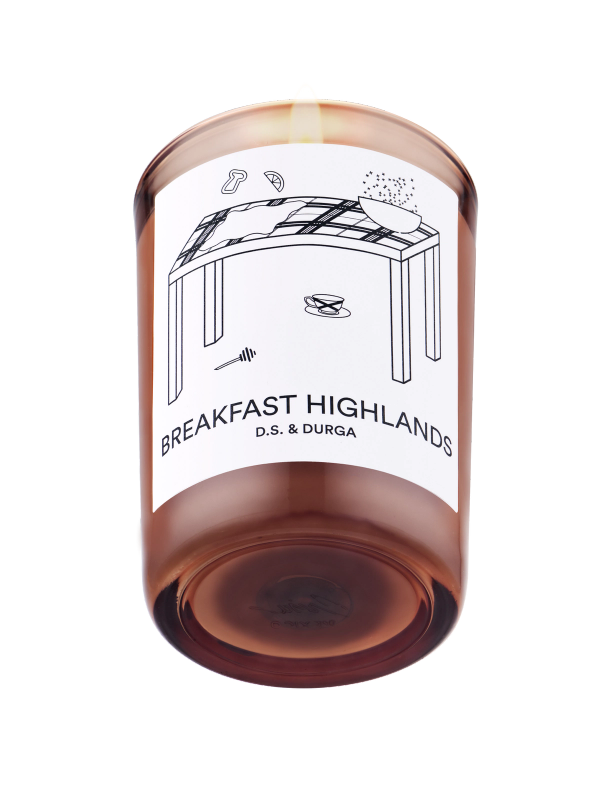 A lit scented candle with a label that reads "breakfast highlands" by d.s. &amp; durga, featuring a simple illustration of a table with a coffee cup and a newspaper.